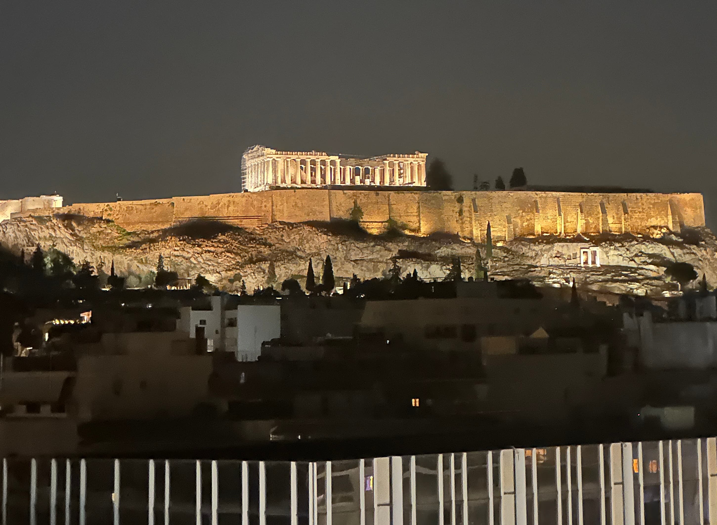 The Acropolis lit up at night above the rooftops of Athens, with the Parthenon at the top and scaffolding on one end of it