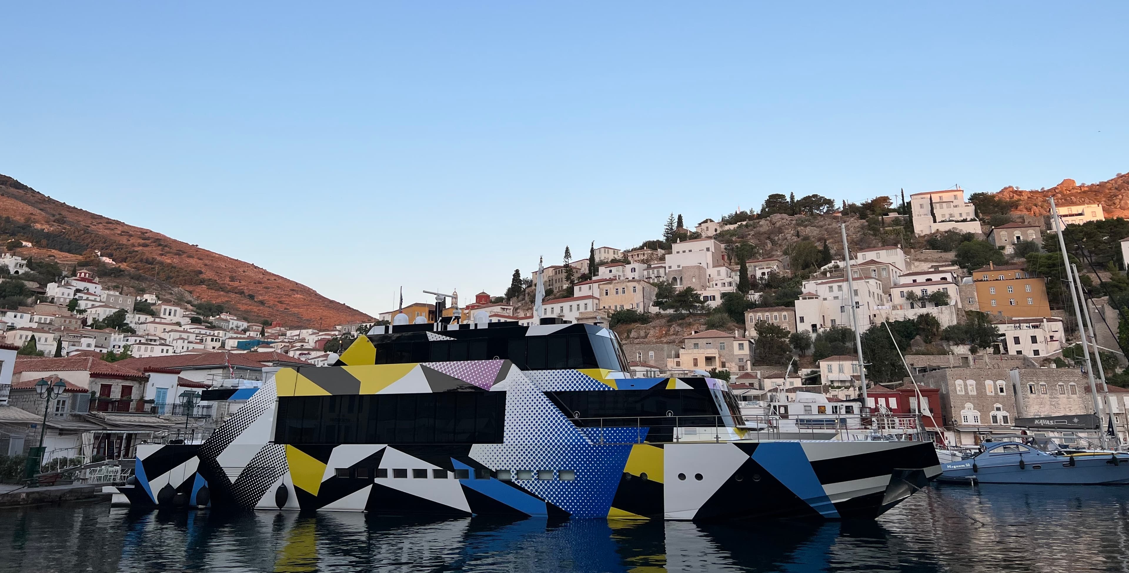 Motor yacht painted in a black, white, yellow and blue geometric pattern, moored in a harbour below a hillside of white and stone houses lit by early evening sun.
