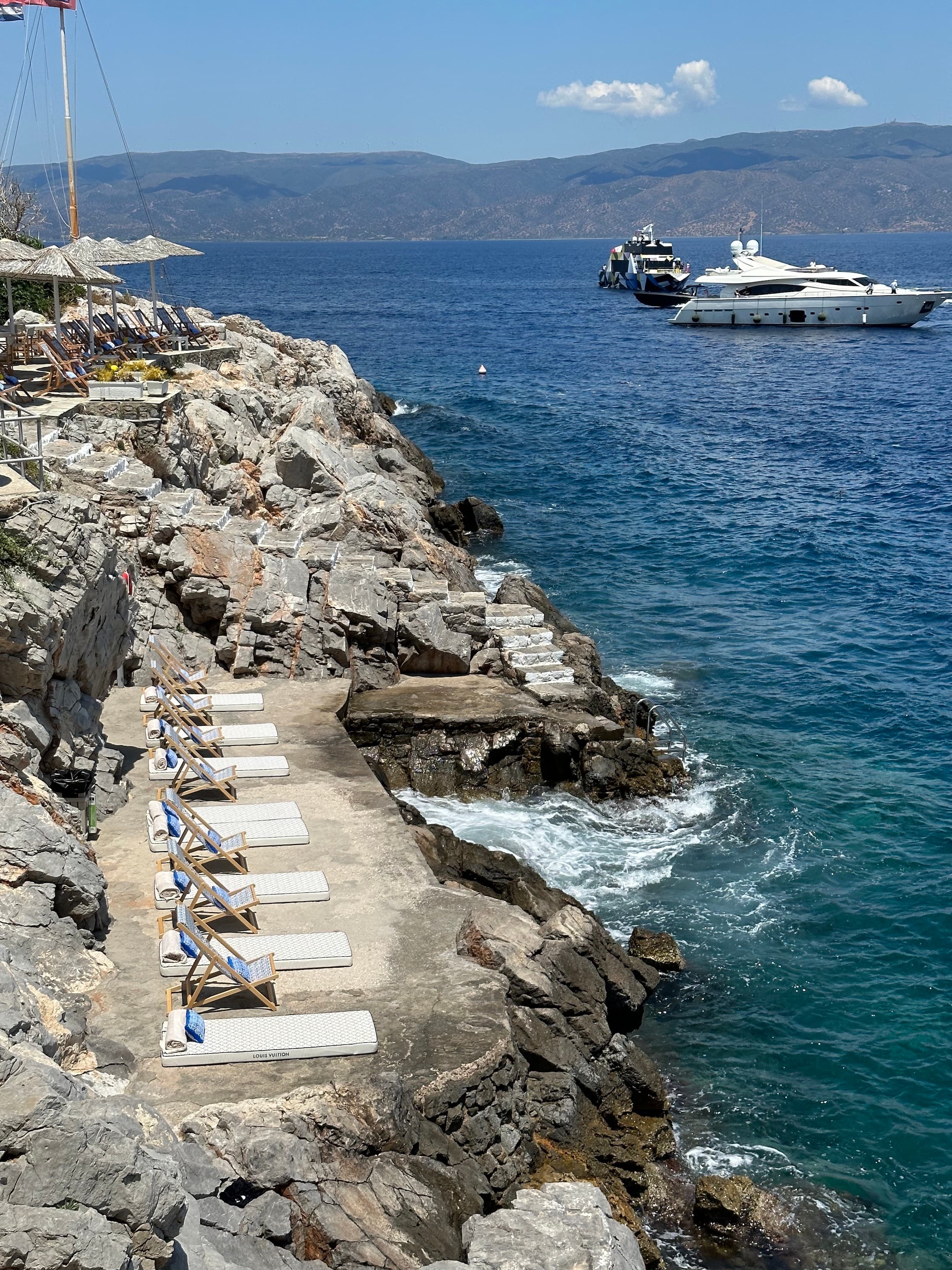 Sun loungers on a stone terrace cut into the rocks on Hydra, with blue sea and two boats anchored offshore