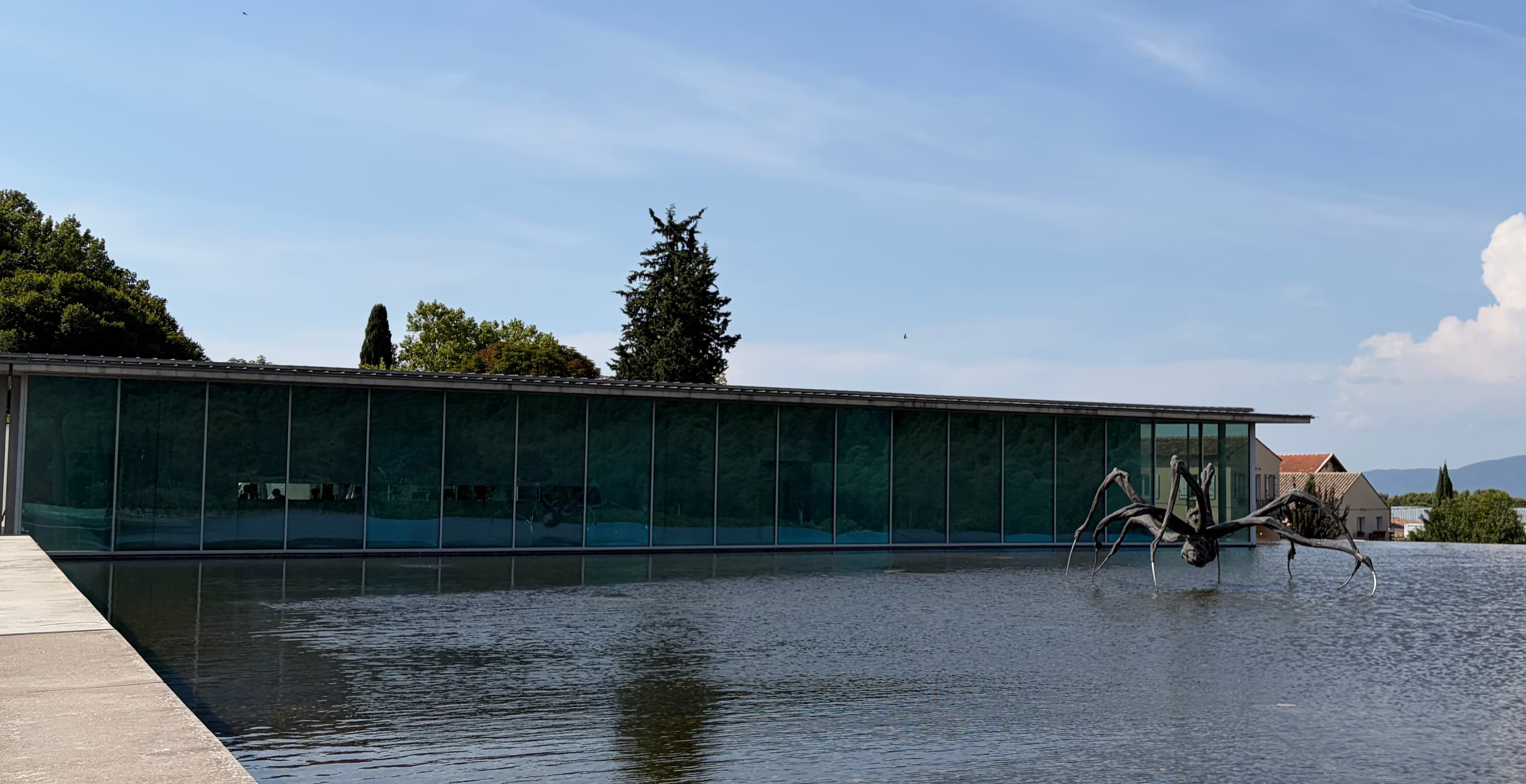 Large dark metal spider sculpture standing in a shallow pool in front of a long glass building at Château La Coste.