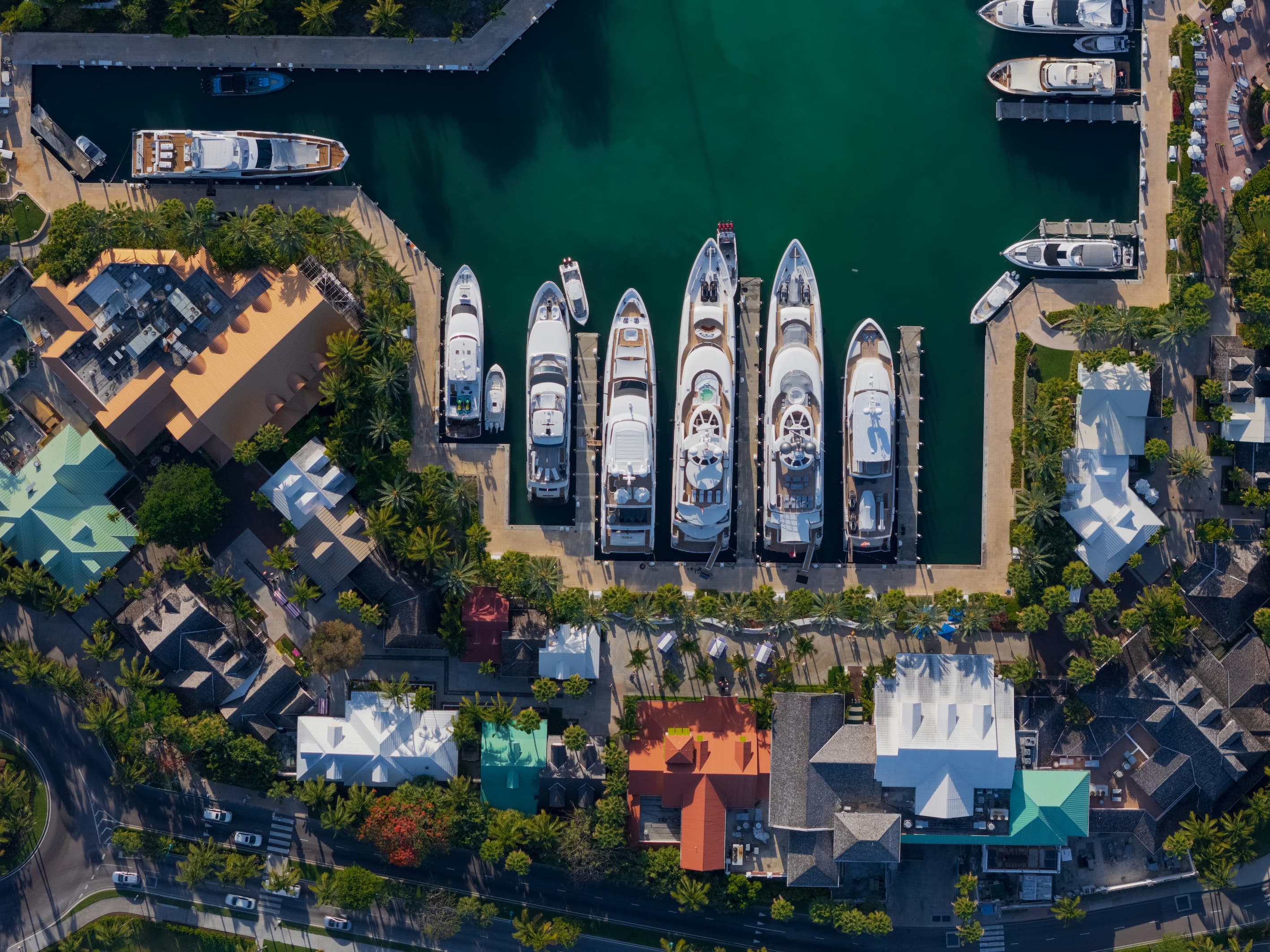 Aerial view looking straight down on a row of superyachts moored side by side in a marina in the Bahamas, with palm trees and colourful roofs along the quay