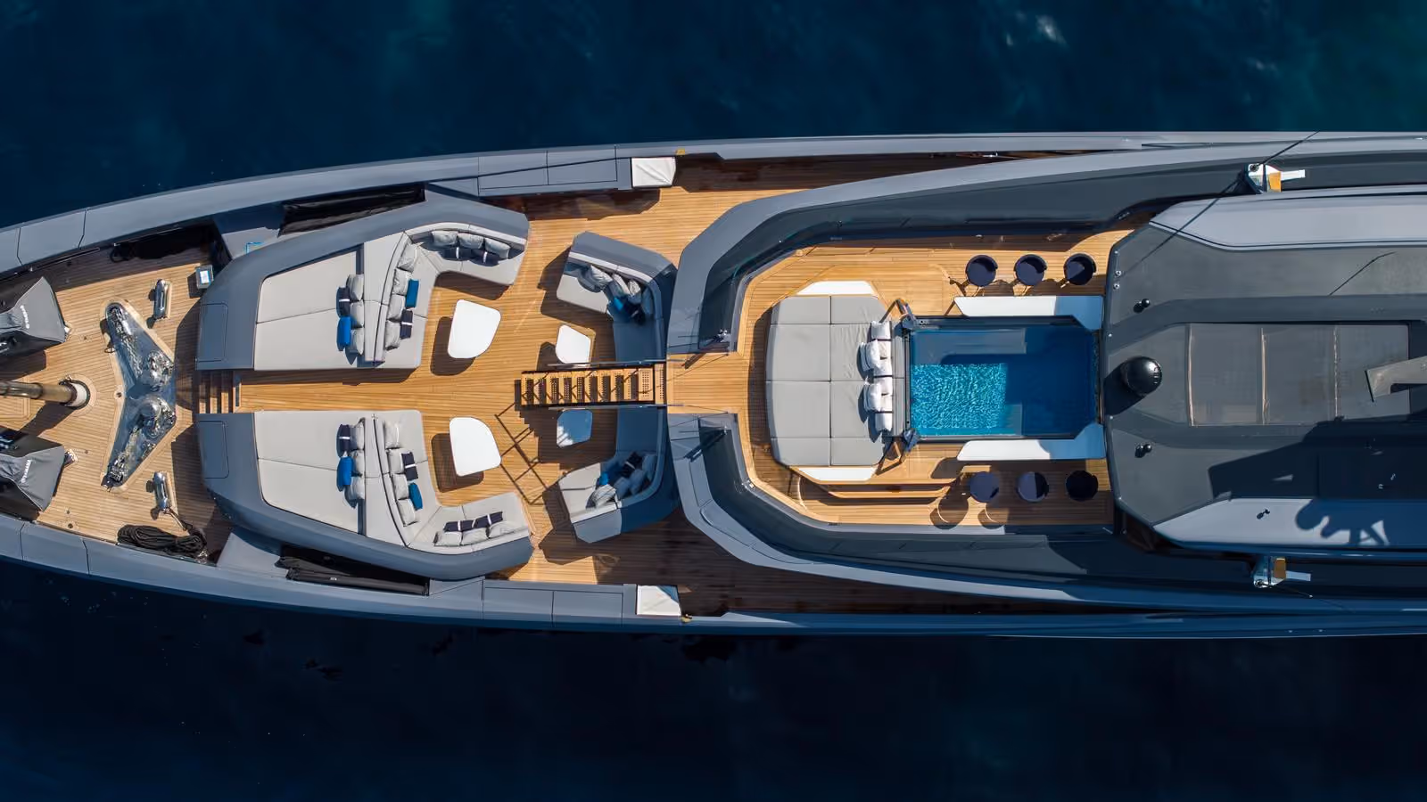 Aerial view of AKULA’s grey hull and teak decks, with lounge seating on the foredeck and a pool surrounded by bar stools.