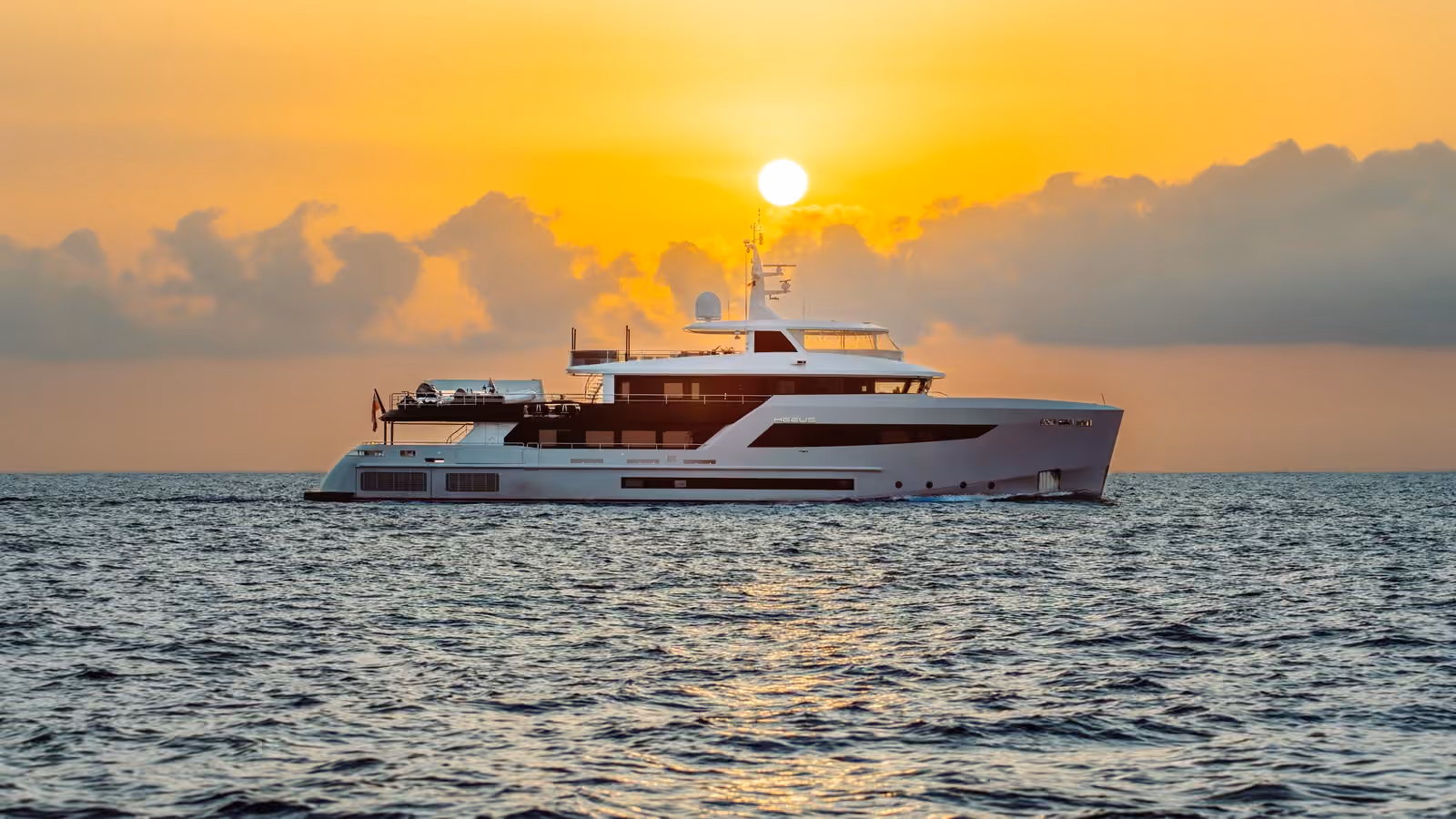 HEEUS, a white motor yacht, cruising on open sea with the sun setting behind her through orange clouds.
