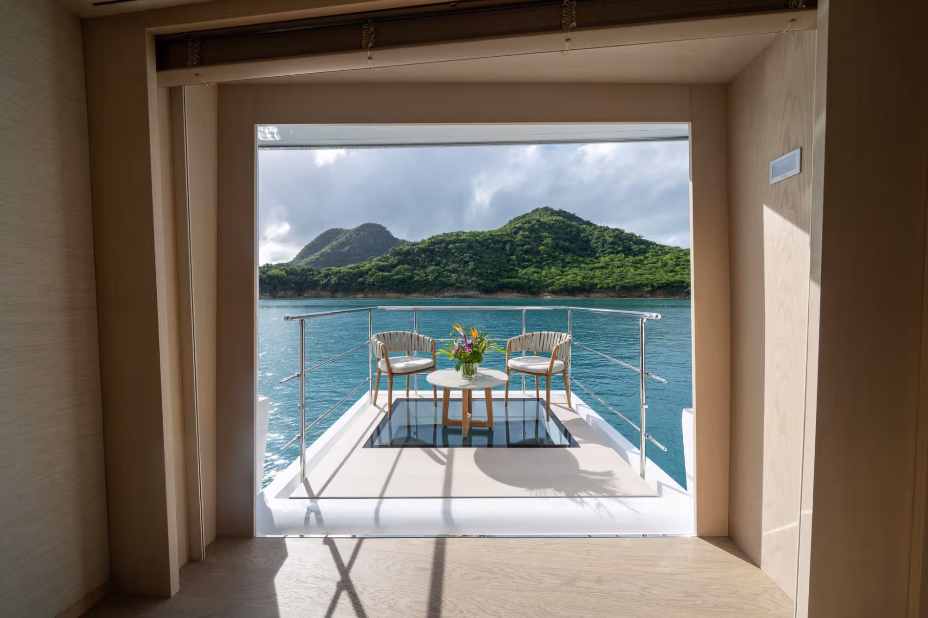 View from WHITE STAR’s wood-panelled interior onto her aft deck, with two chairs, a small table with tropical flowers and a glass floor panel, facing turquoise water and green hills.