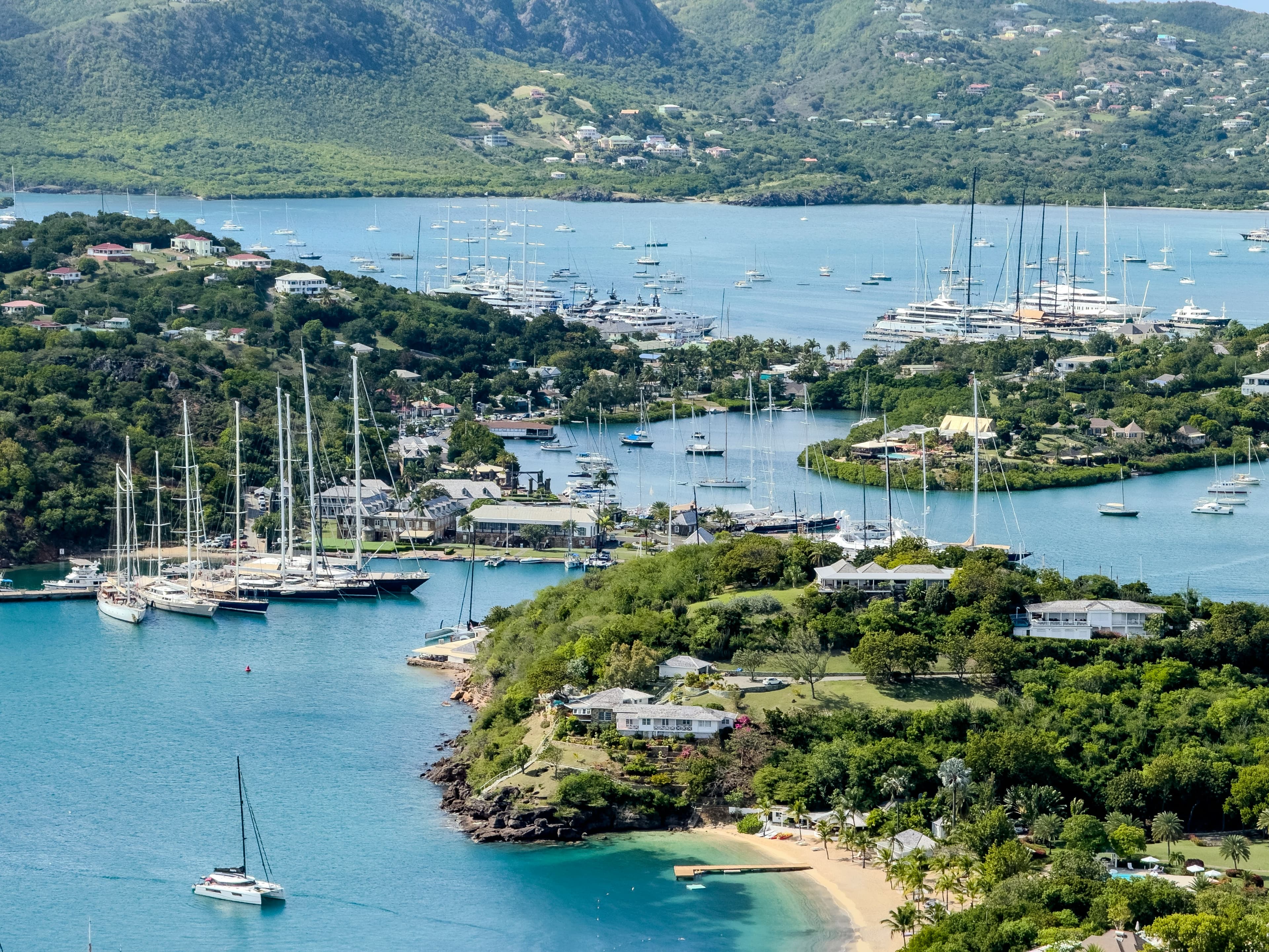 Superyachts moored in English Harbour and Falmouth Harbour, Antigua, seen from the hills above, with Nelson’s Dockyard and a beach in the foreground.