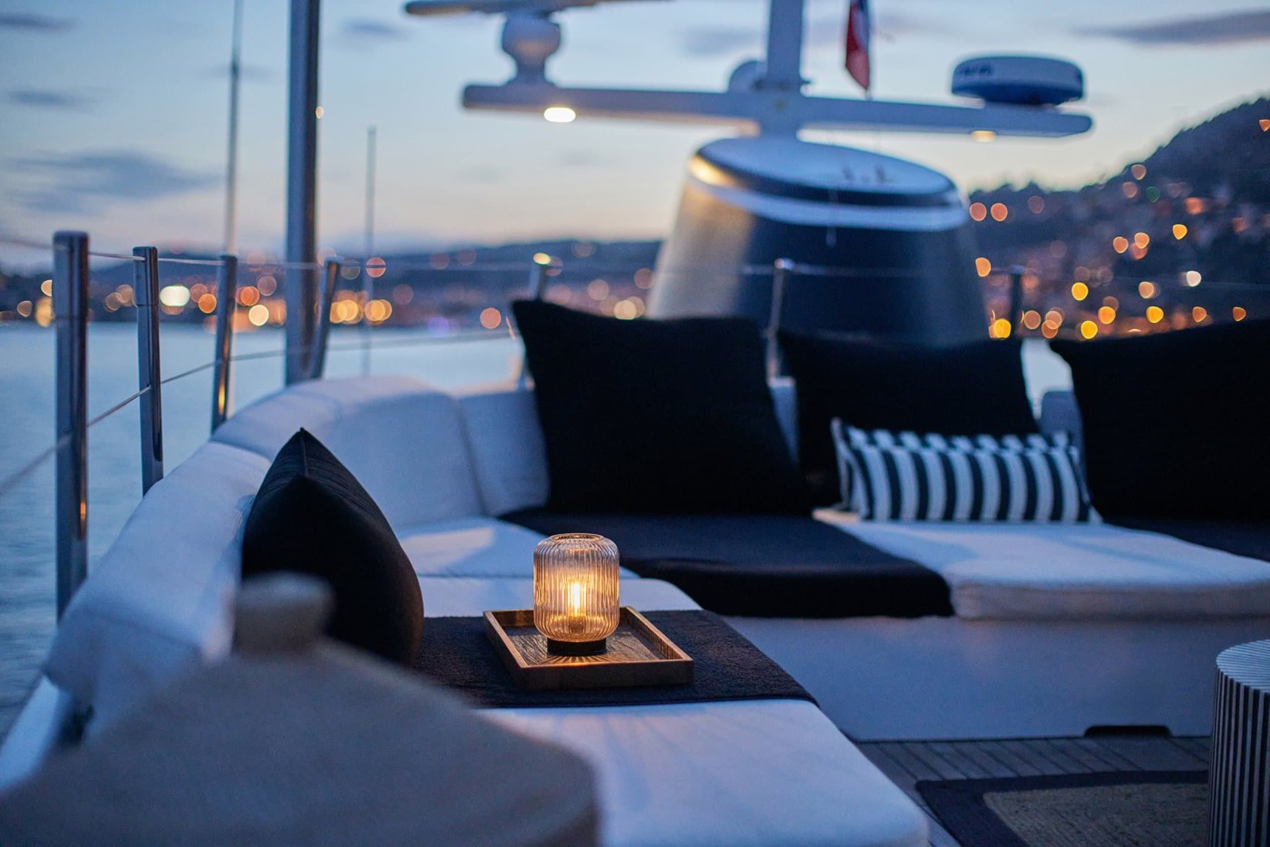 SOUNION II - deck seating with a lit candle and cushions at dusk with harbour lights in the background | Drivas Yachts